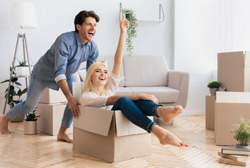 Home Owners. Couple Having Fun In New House. Man Riding Woman In Moving Box Celebrating Buying Own Apartment. Copy Space