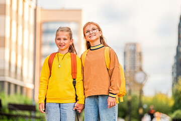 Two happy children walk to school with backpacks along a sunny city street.