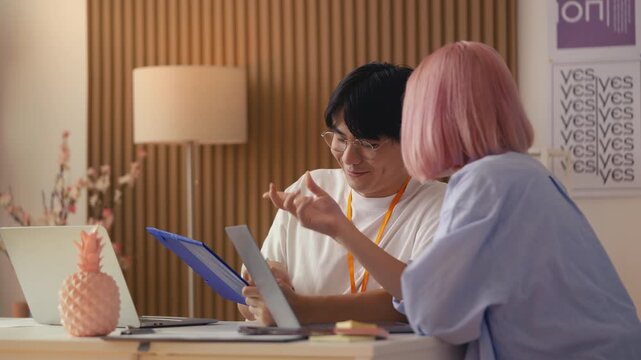 Pink-haired web designer and her Asian colleague discuss work task in office