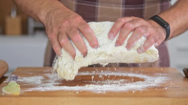 Kneading pizza dough by hand on a wooden board. A man works in a home kitchen, preparing a family meal. Natural light illuminates the authentic kitchen setting.