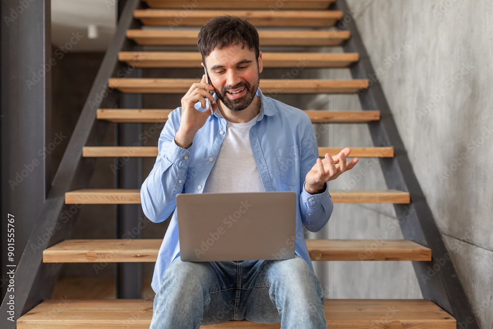 Poster a man is sitting on wooden stairs in a home, talking on a cell phone while holding a laptop on his l