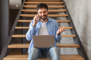 A man is sitting on wooden stairs in a home, talking on a cell phone while holding a laptop on his...