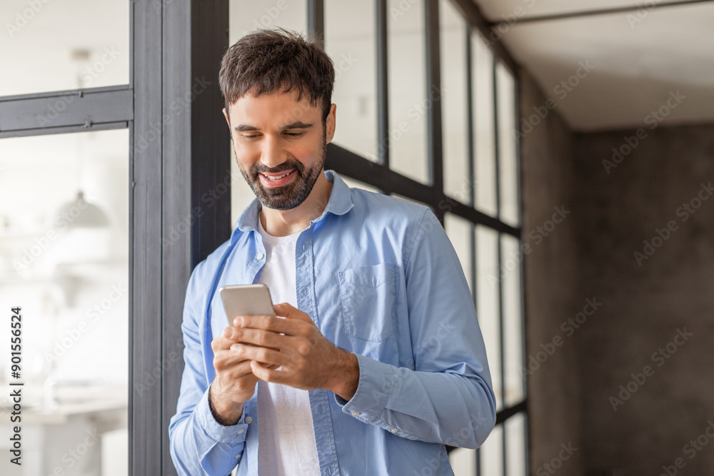 Aufkleber a man with a beard stands near a window in a modern office setting, smiling as he uses his smartphon