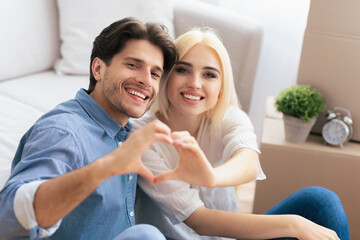 A couple sits together in their new home, smiling and making a heart shape with their hands.