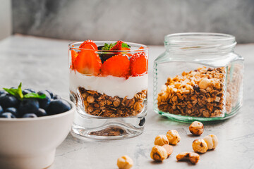 Healthy breakfast, parfait dessert with granola, Greek yogurt and strawberries and blueberries in a transparent glass on a gray background with ingredients. Selected focus