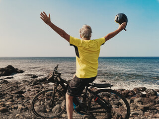 Rear view of senior sporty man stops after a bike ride on the beach looking at horizon over sea...