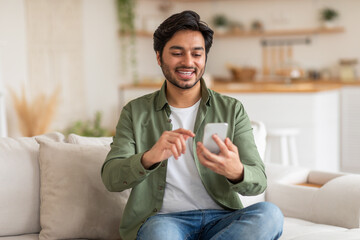Arab young man, wearing a green shirt and jeans, smiles while looking at his phone. He is sitting...