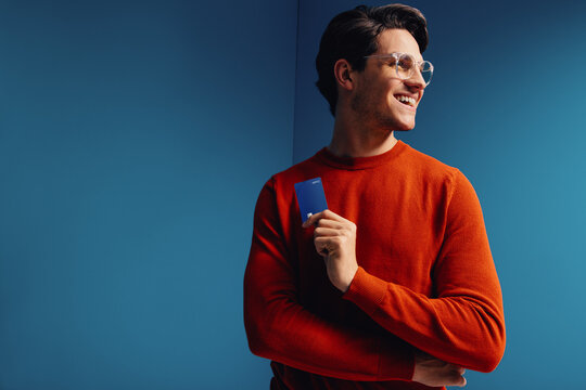 Smiling young man holding a credit card in a vibrant, colorblocked red and blue setting, showcasing wealth and modern finance