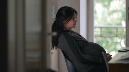 Pregnant woman sitting at a desk, side view, thoughtful expression, indoor setting, natural light from window, expecting mother, contemplation, peaceful moment, home environment