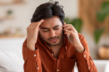 Arab man with dark hair and a beard is on a phone call inside. He has a worried expression and is...