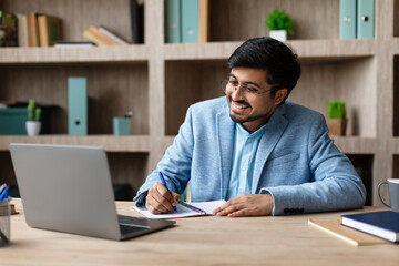 A smiling Indian man in a blue suit is sitting at a desk, taking notes in a notebook while looking at his laptop. The man is focused on his work and appears happy.