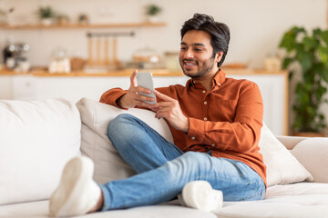 A young Arab man in a casual orange shirt and blue jeans is sitting on a white couch and using his...