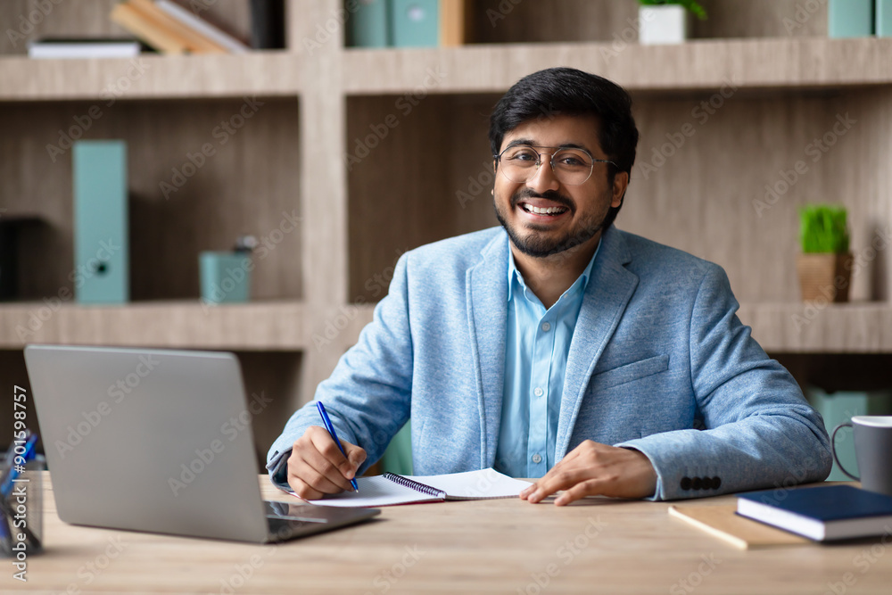 Aufkleber a smiling indian man with a beard and glasses sits at a desk in a modern office, working on a notebo