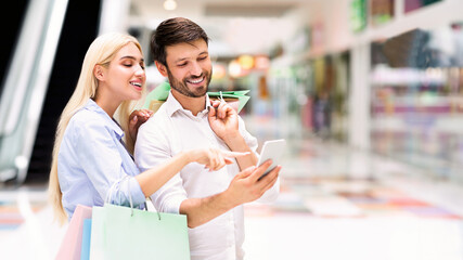 A couple is shopping together at the mall. They are looking at a smartphone, smiling and appearing to be happy. The woman is holding shopping bags and pointing at the screen of the smartphone