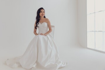 Fototapeta premium A beautiful bride in a white wedding dress sits in a chair in a white room. She looks to the side with a smile on her face.