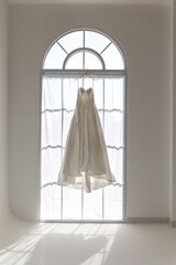 A white wedding dress hangs in front of a large window, bathed in soft sunlight. The dress is a classic A-line silhouette with a sweetheart neckline.
