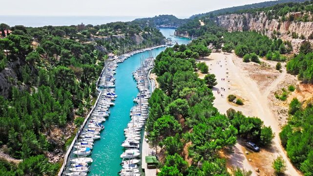 Aerial view of a long, narrow inlet featuring a marina lined with sailboats and hiking path with scenic water views. Calanque de Port-Miou in the eastern calanque between Marseille and Cassis.
