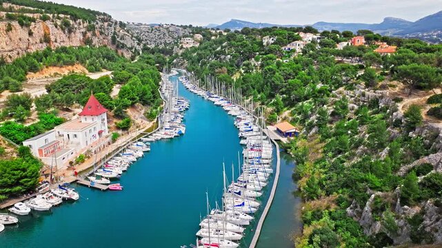 Aerial view of a long, narrow inlet featuring a marina lined with sailboats and hiking path with scenic water views. Calanque de Port-Miou in the eastern calanque between Marseille and Cassis.
