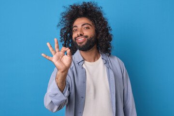 A cheerful individual with curly hair gives an enthusiastic okay sign while smiling against a vibrant blue backdrop.