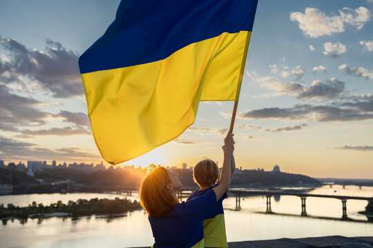 Mom and son with a large Ukrainian flag stand high on the roof of a house in front of the Dnieper River in Kyiv and sunset. Patriotism, drawing attention to the war in Ukraine. Independence Day