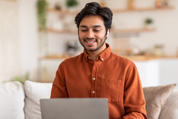 Arab man sits on a couch in his home, smiling while looking down at his open laptop. He is wearing earbuds and an orange button-down shirt.