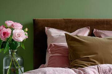 A close-up shot of the headboard, showcasing soft pink and rich brown velvet cushions on an elegant bed against a green wall background. A vase with delicate roses adds to the romantic ambiance.