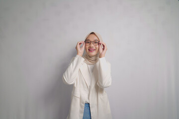 A smiling young woman wearing a white blazer and hijab, adjusting her glasses