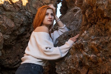 Close up portrait of a red-haired beautiful young girl with long loose hair, who stands leaning on a rock on an autumn beach, dressed in a white sweater