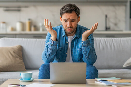 A man sits on a couch in his living room, looking confused as he stares at his laptop. His arms are raised in a gesture of frustration. A cup of coffee sits on a table in front of him.