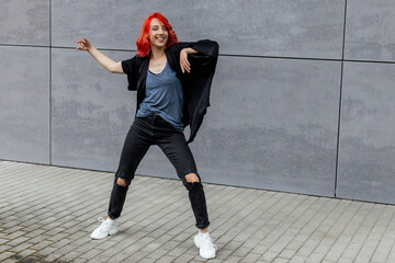 Happy positive emotional dancer curling in dance. Woman with red hair dancing on urban street, on gray wall background. International dance day.