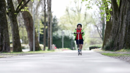Fototapeta premium Young child wearing helmet and green jacket riding a scooter on a park path, enjoying outdoor activity, safety gear, vibrant spring day, active lifestyle, childhood fun