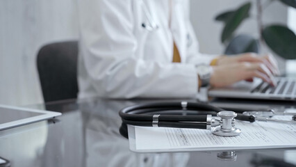 Close-up of doctor's desk with stethoscope and clipboard. Physician is using a laptop computer at the background. Medicine concept