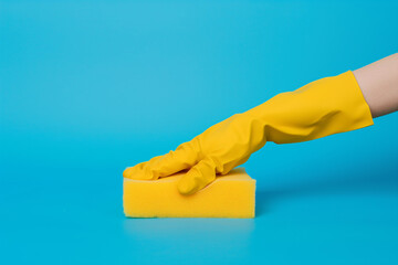 A woman's hand in a yellow glove holds a yellow cleaning sponge on a blue background. Photo concept...