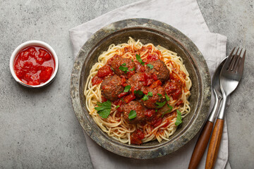 Spaghetti and meatballs with tomato sauce served on rustic plate with cutlery and tomato sauce on the side, on a gray background