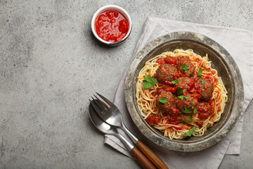 Spaghetti and meatballs with tomato sauce served on rustic plate with cutlery and tomato sauce on the side, on a gray background copy space