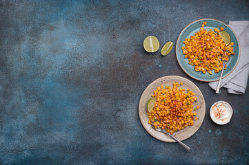 Two plates of esquites, a popular mexican street food dish, are served with lime wedges and sour cream with chili powder
