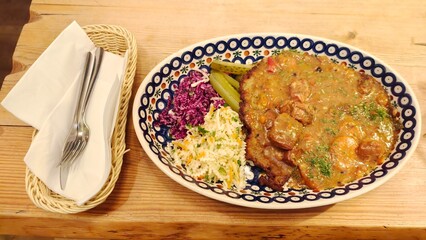 On the wooden table there is a ceramic plate with fried potato pancake and meat goulash, coleslaw and pickles. Nearby is a wicker basket with metal knives and forks on napkins. Dinner time