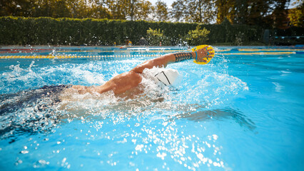 Wearing yellow paddles and white cap, athletic man, swimmer practices his freestyle technique in outdoor pool under sun. Concept of professional sport, competition, active and healthy lifestyle. Ad