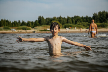 Fototapeta premium A smiling boy with arms outstretched stands in the water at a beach. Other beachgoers are visible in the background under a clear sky