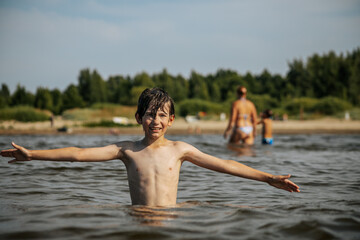 Fototapeta premium A smiling boy with arms outstretched stands in the water at a beach. Other beachgoers are visible in the background under a clear sky