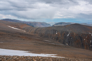 Dramatic top view from pass above large sharp ridge to alpine deep valley among big cliffs far away in rain under cloudy sky. Bleak atmospheric landscape with high rocky mountains under rainy clouds.