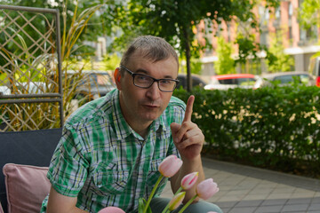  A man sits at an outdoor table with pink tulips, raising a finger as if making a point.