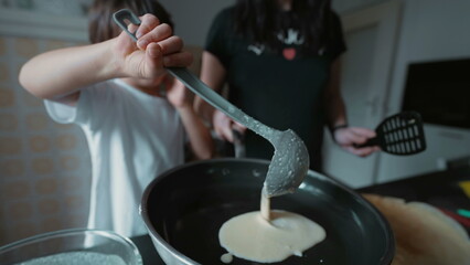 Child learning to pour pancake batter into pan, kitchen setting, focused expression, parent supervision, home environment, morning light, family activity, white t-shirt, making breakfast