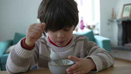 Young boy enjoys a bowl of yogurt at the table, lifting the spoon to his mouth with a thoughtful expression, capturing a serene and candid moment in a home environment