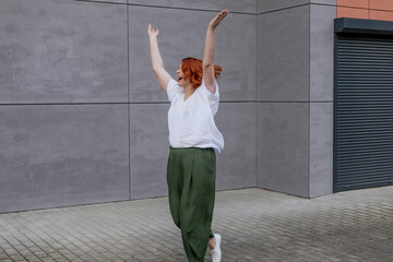 Happy positive dancer curling in dance. Woman with red hair dancing on urban street, on gray wall...