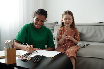 Happy teenage sitter and cute little girl with crayons drawing together in living room while one of them sitting on grey couch