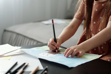 Cropped shot of little girl with crayon drawing picture of her pet while sitting by table with paper sheets and many colorful pencils