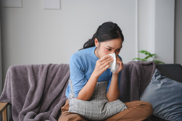 Sick young woman blowing her nose with a paper tissue while sitting on the sofa at home