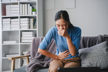 Young woman sitting on a sofa and feeling sick, she is covering her mouth with her hand and holding...