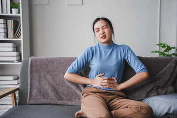 Young woman is sitting on her sofa, hunched over in pain and holding her stomach